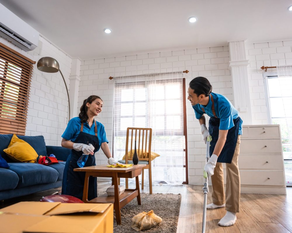 Asian young man and woman cleaning service worker work in living room. Attractive housekeeper cleaner team wear apron and cleaning messy dirty floor for housekeeping housework and chores in house.